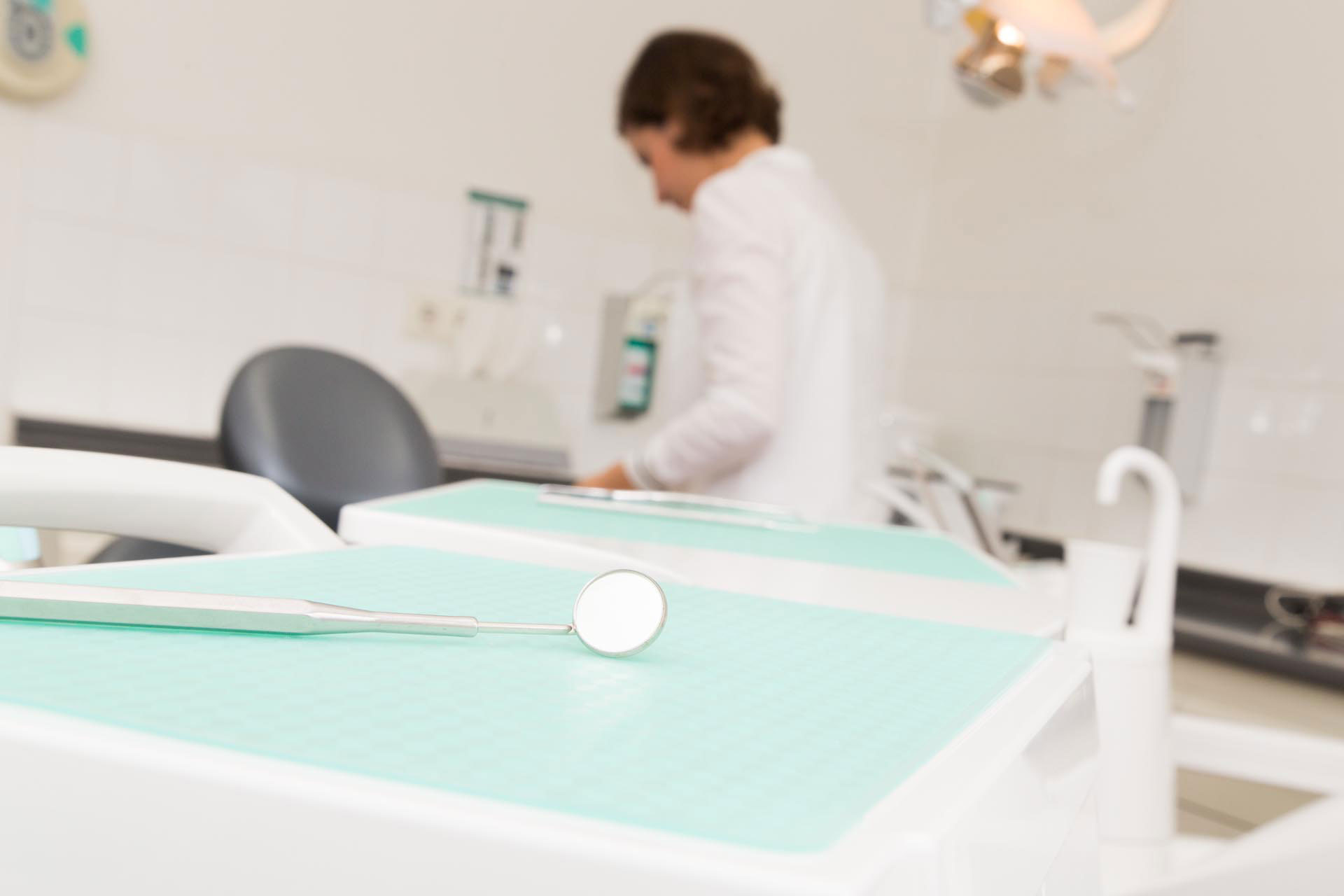 Zahnarztspiegel im Fokus auf einer Ablage für zahnärztliche Instrumente in einem Praxisraum, mit Frau Dr. Sarah Herrmann unscharf im Hintergrund.